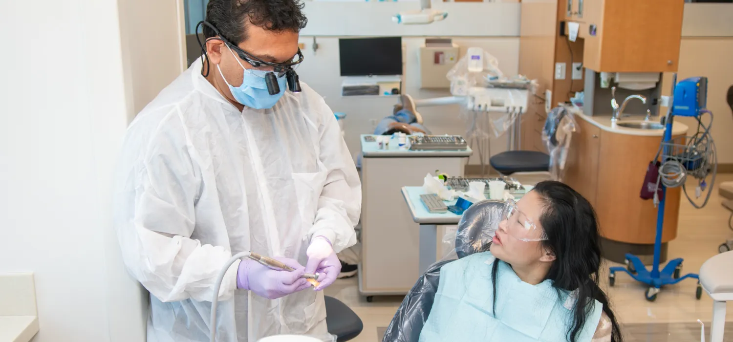 A student prepares instruments while a patient seated in a dental chair looks on in a dental clinic