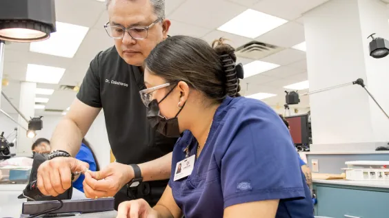 A dental instructor guides a student working at a lab bench in a dental school clinic