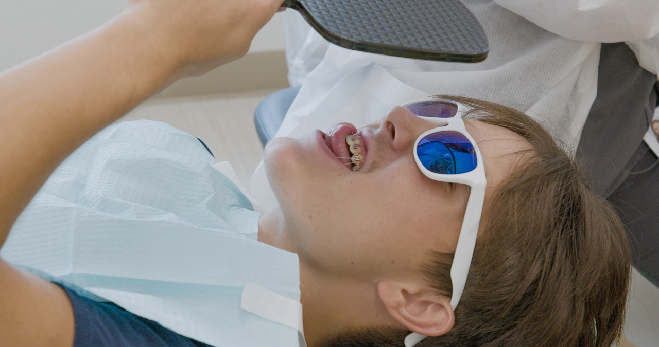 A young patient with braces reclines in a dental chair wearing white sunglasses holding a mirror to see their braces