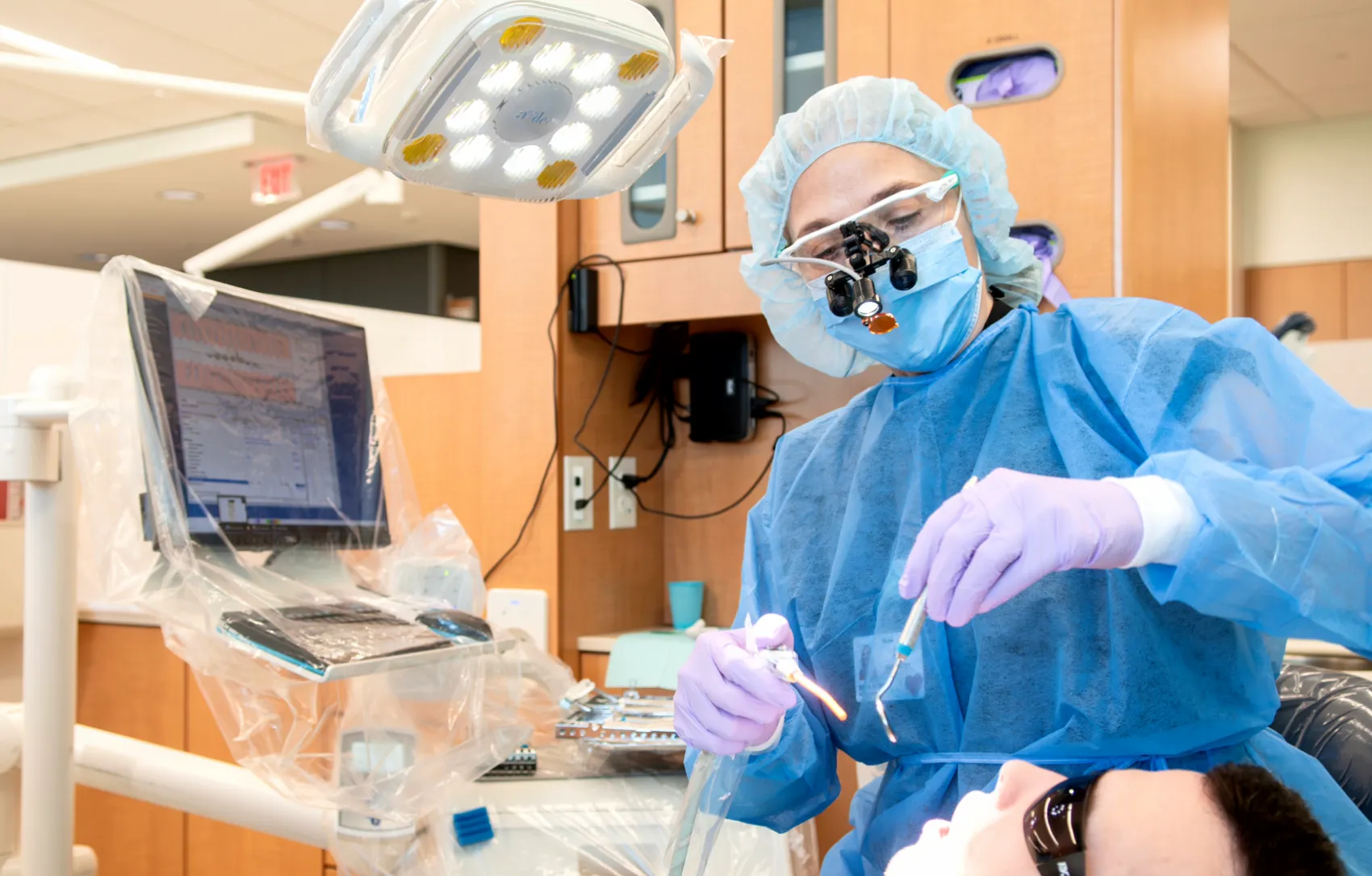 A dental surgeon in full surgical attire performs a procedure under an operating light.